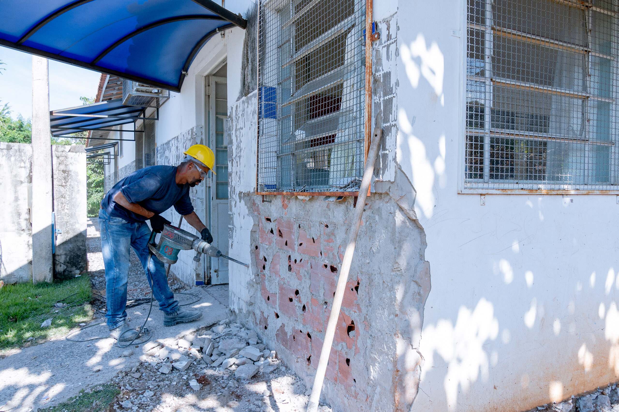 A imagem mostra um trabalhador da construção civil usando um martelo pneumático para demolir uma parede externa de um prédio. O trabalhador está usando um capacete amarelo e óculos de segurança. Ele está usando jeans e uma camisa azul escura. A parede é de tijolo e reboco, e partes dela estão quebradas e descascadas. O prédio parece antigo e está em processo de reforma ou demolição. Há uma cobertura azul acima da área onde o trabalhador está trabalhando, protegendo-o do sol. No chão há destroços de tijolos e concreto quebrados. Ao fundo, há um pouco de vegetação e um pedaço de muro. A cena está iluminada pelo sol. A imagem sugere um trabalho manual intenso em condições externas quentes.