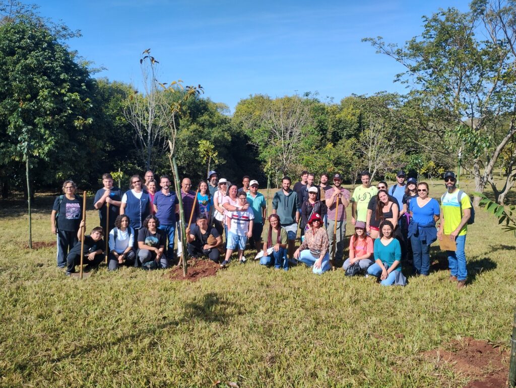 A imagem mostra um grupo de pessoas reunidas ao ar livre em uma área gramada com árvores ao fundo. O grupo parece estar participando de uma atividade de plantio de árvores, pois há mudas recém-plantadas no solo e algumas pessoas estão segurando pás.