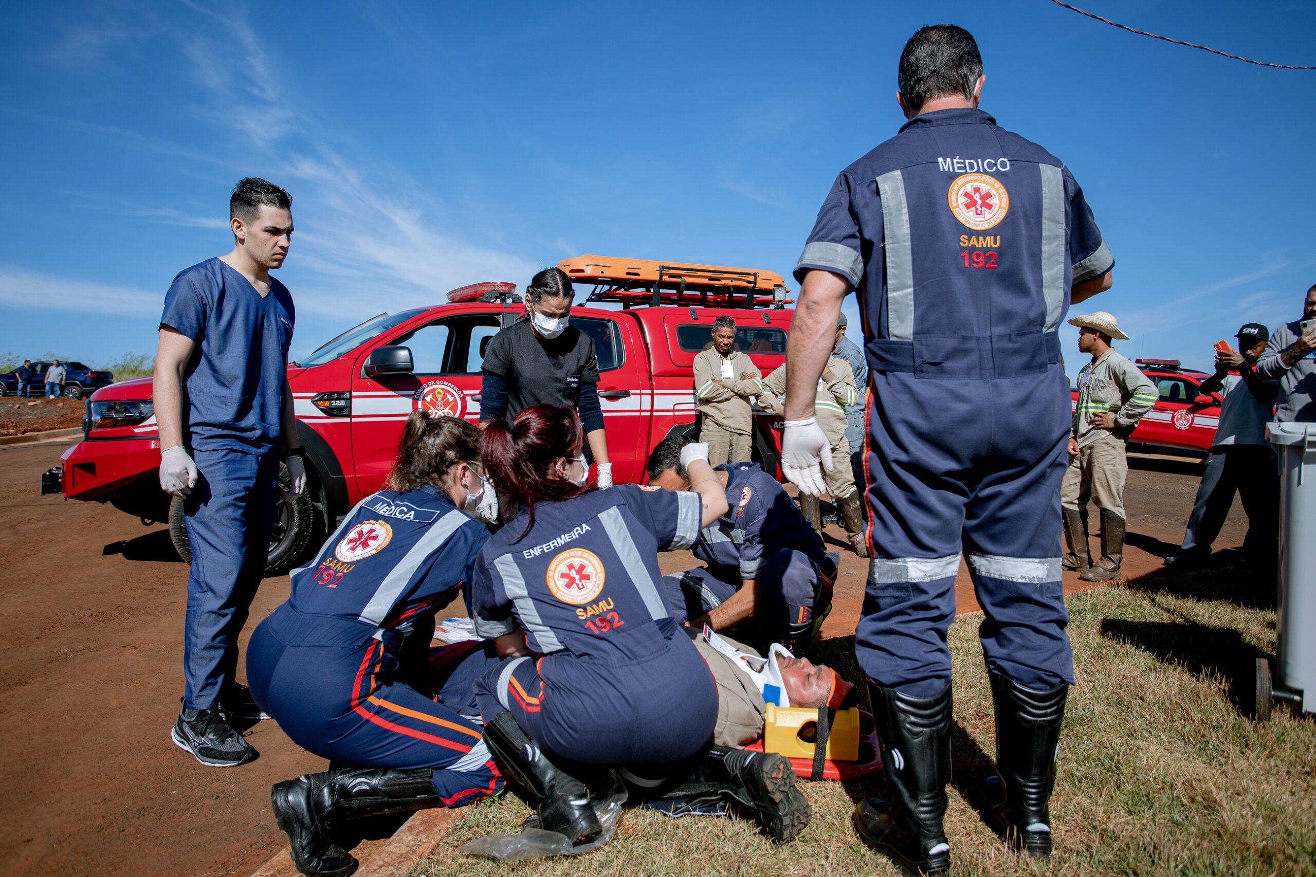 A imagem mostra uma cena de atendimento de emergência médica em uma área externa, aparentemente rural. O foco principal é um grupo de profissionais de saúde do SAMU (Serviço de Atendimento Móvel de Urgência) atendendo a uma pessoa deitada em uma maca improvisada. Há vários profissionais envolvidos: um médico, enfermeiros e técnicos, todos uniformizados com a identificação do SAMU. Eles estão prestando os primeiros socorros à vítima, que parece estar inconsciente ou ferida. Ao fundo, há uma caminhonete vermelha do Corpo de Bombeiros, sugerindo a participação deles na ocorrência. Há também outras pessoas presentes, algumas assistindo à cena e outras aparentemente envolvidas no atendimento, mas não uniformizadas como os profissionais de saúde. O cenário é de dia, com céu azul e vegetação esparsa ao redor. A atmosfera é de urgência e preocupação.