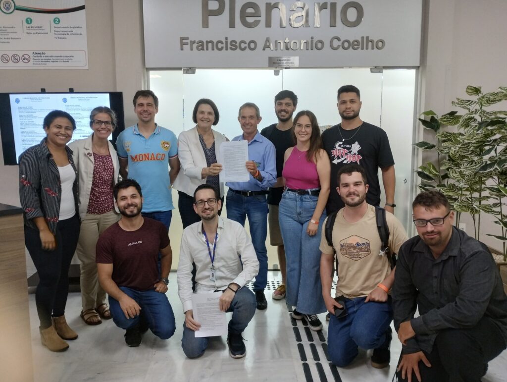Nancy Thame, da Secretaria Municipal de Agricultura e Abastecimento (Sema), aparece segurando o diploma da Câmara, ao lado do agricultor Vanderlei. Em pé estão 8 pessoas e agachados quatro jovens.