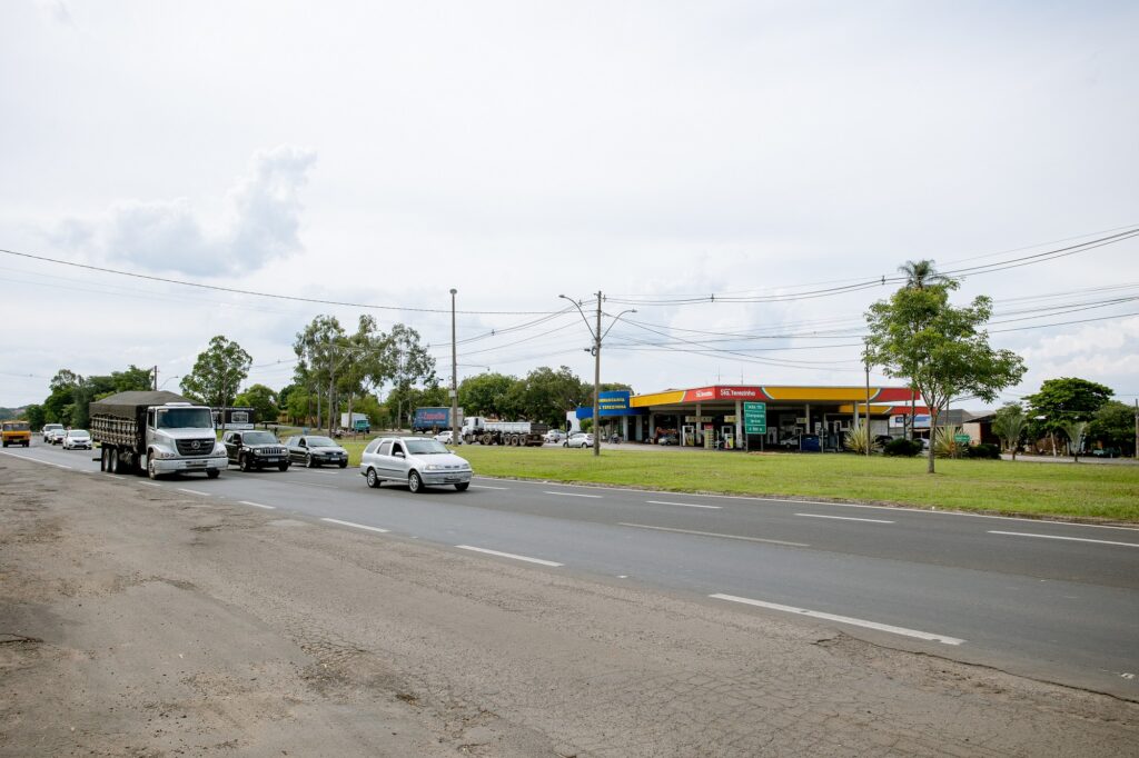 A imagem mostra uma estrada movimentada com vários veículos, incluindo carros e um caminhão, trafegando em ambas as direções. No lado direito da imagem, há um posto de gasolina com um telhado amarelo e vermelho, e várias placas indicando serviços e preços. O posto de gasolina é cercado por um pouco de vegetação, incluindo árvores e grama. Também há linhas de transmissão de energia visíveis acima da estrada. O céu está nublado, sugerindo um clima encoberto.