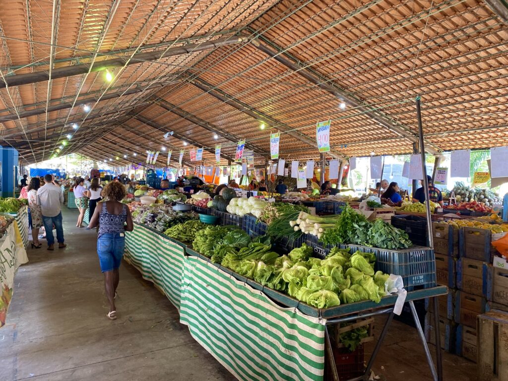 Bancas de verduras e legumes colocados em barracas de madeira. Ao lado algumas pessoas observar os produtos, entre eles alface, almeirão, couve, beringela, etc