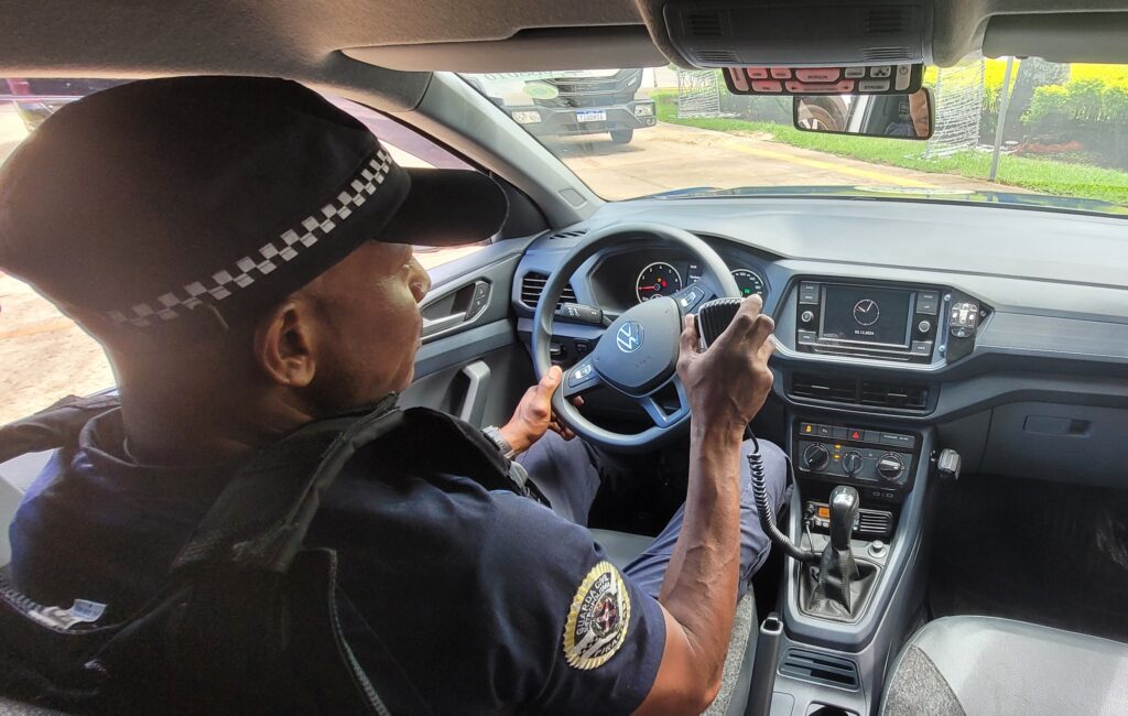 A imagem mostra um policial sentado ao volante de um carro da polícia Volkswagen. Ele está usando um uniforme e um boné de polícia. Ele está segurando um rádio e parece estar transmitindo. O interior do carro é cinza e moderno. A imagem está bem iluminada e o foco está no policial.