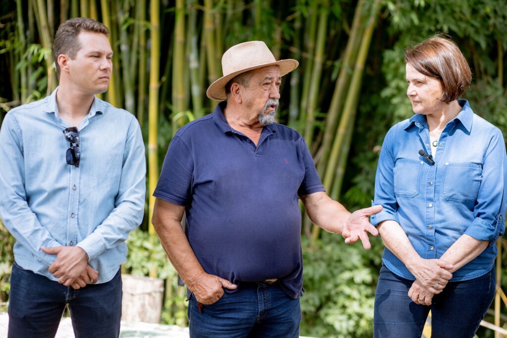 Agricultou de chapéu e camiseta azul marinho e calça jeans escura, gesticula para a secretária muniicpal de agricultura, Nancy Thame. E do seu lado direito está um jovem de camisa azul clara e calça jeans escura, com óculos de sol preto pendurado na camisa.