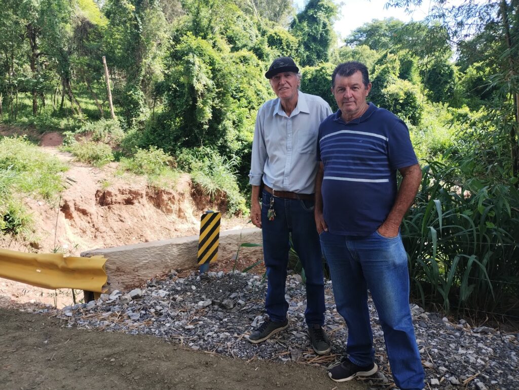 Dois agricultores, sendo um de boné escuro na cabeça, camisa clara e calça jeans, com chaves penduradas no cinto marron, ao lado de outro agricultor, com mãos no bolso, de camiseta azul e listras brancas e calça jeans clara.