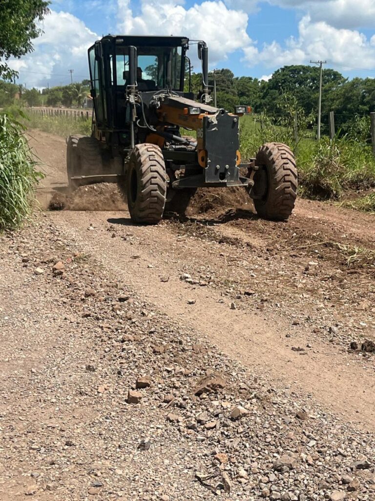 Trator com quatro rodas aplaina estrada rural, tendo ao lado vegetação.