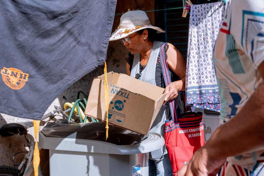 Mulher de chapéu branco com detalhes emamarelo, de óculos, segura uma caixa de papelão, tendo a tiracolo uma sacola vermelha comdetalhes em azul escuro. Ela observa materiais encontrados em cima de um tanquinho branco.