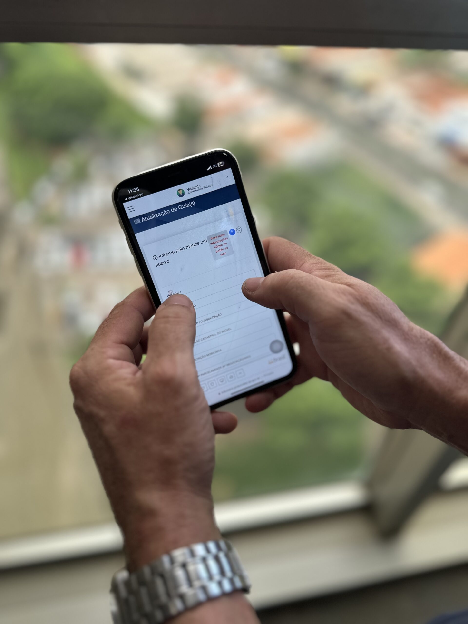 A imagem mostra um par de mãos masculinas segurando um smartphone. O telefone exibe uma tela aberta no sistema tributário da prefeitura. Ao fundo, através da janela, é possível ver uma vista urbana, com edifícios e uma estrada. A luz natural entra pela janela, iluminando suavemente a cena.