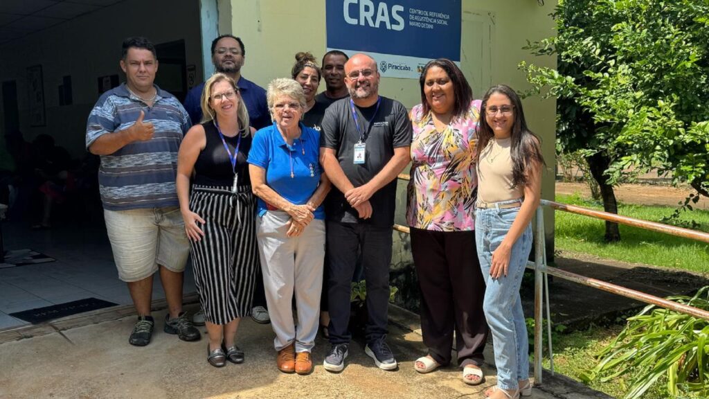 Ao centro posando para fotos está Adriano Camargo, calvo, de óculos e crachá e camiseta e tênis escuros e tenis cinza, rodeado por líderes comunitários do Mário Dedini, em frente ao CRAS Mário Dedini