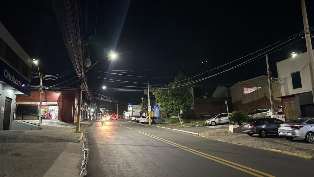 Vista noturna de Avenida extensa,com iluminação Led. Do lado direito aparecem diversos carros estacionados. As lâmpadas refletem a iluminação nova .