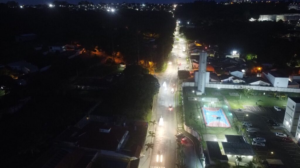 Vista noturna de avenida com nova iluminação.Do alto se vê condomínios e vegetação verde ao redor.