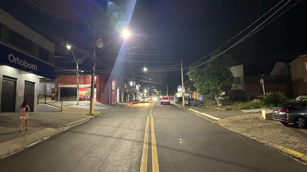 Avenida a noite iluminada. Ao lado se vê uma mulher de blusinha preta e short branco com mochila nas costas, caminhando pela calçada. Na avenida se vê carros e ao lado alguns carros estacionados.
