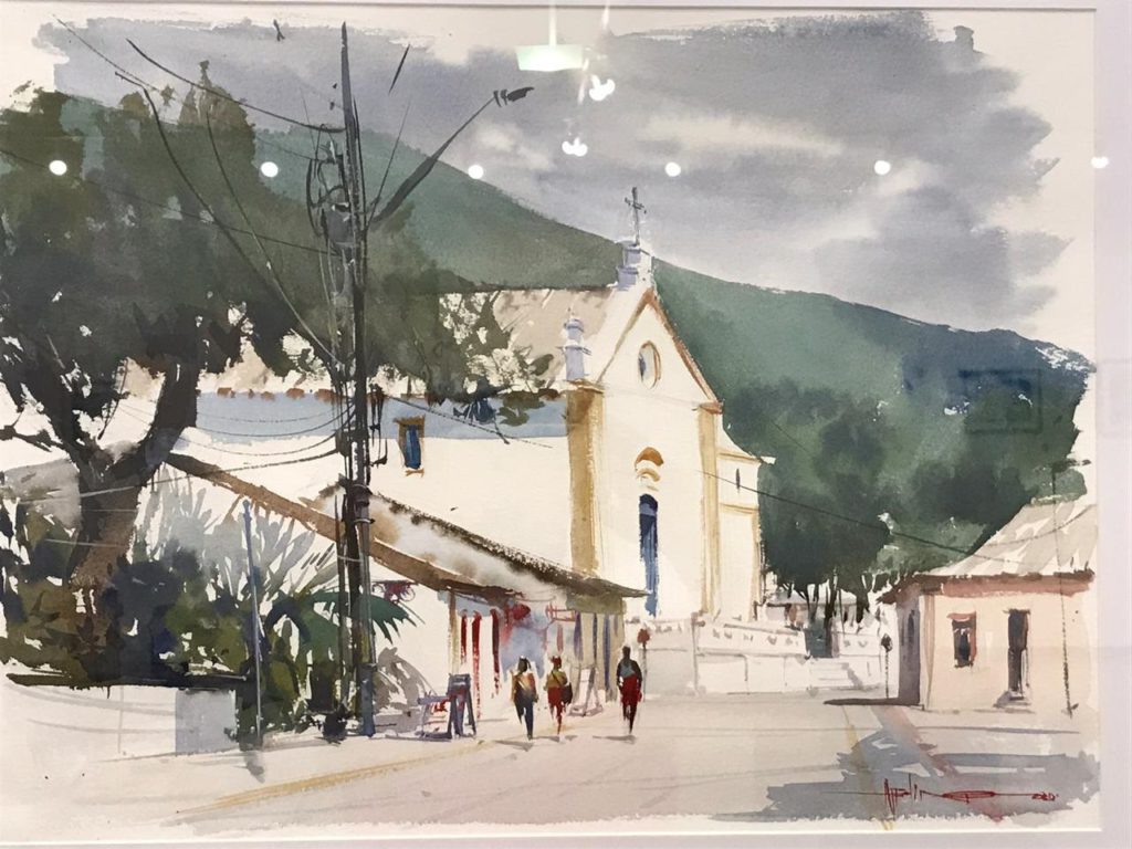 Aquarela retratando uma cena urbana com uma igreja de fachada clara e detalhes em tons de amarelo ao centro, cercada por edificações simples e vegetação exuberante. Ao fundo, colinas verdes contrastam com o céu nublado em tons de cinza. Um poste com fios elétricos corta a cena, adicionando profundidade. No primeiro plano, três pessoas caminham tranquilamente pela rua, trazendo um toque de vida à composição. A obra transmite uma atmosfera serena e bucólica.