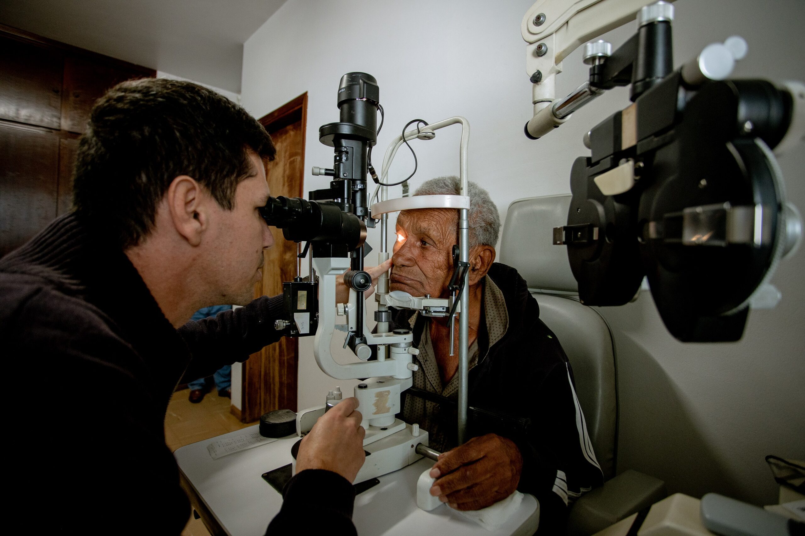 Uma imagem mostra um homem mais velho com cabelos grisalhos sentado numa cadeira de exame de olhos, enquanto um homem mais jovem, de cabelos escuros, olha através de um oftalmoscópio para examinar os olhos do homem mais velho. O homem mais velho parece estar calmo e cooperativo, enquanto o homem mais jovem parece estar concentrado e profissional. O ambiente parece ser um consultório médico ou clínica, com paredes claras e equipamento médico visível. A iluminação é suave e difusa, sem sombras duras. As cores são principalmente neutras, com tons de cinza, bege e preto. A textura da roupa dos homens é visível, e os detalhes do equipamento médico são nítidos. O foco da imagem é o exame de olhos, e a composição é próxima e íntima. Não há texto visível na imagem. Não há marcas ou logotipos visíveis. A qualidade da imagem é boa, com boa resolução e detalhes. A perspectiva é de um ângulo ligeiramente elevado, olhando para baixo para os homens. Não há elementos fotográficos notáveis. Não há pistas contextuais visíveis. As relações espaciais mostram o homem mais jovem próximo ao homem mais velho, ambos interagindo diretamente com o equipamento médico.