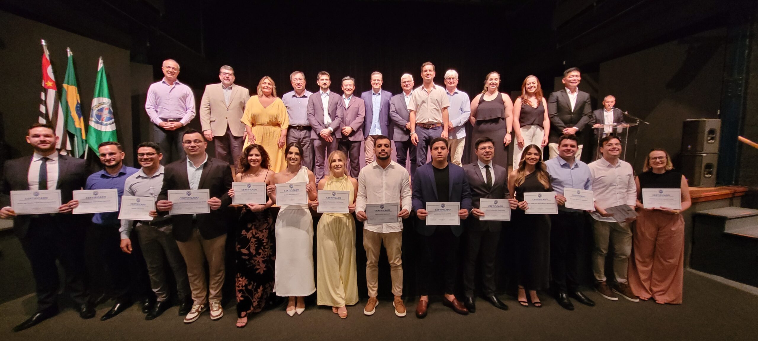 A imagem mostra um grande grupo de pessoas em pé em um palco, em frente a um fundo escuro. Eles estão segurando certificados e parecem estar em algum tipo de cerimônia de formatura ou premiação. Há bandeiras atrás deles, incluindo as bandeiras do Brasil e de outros países. As pessoas estão vestidas de forma semi-formal, com homens usando ternos ou camisas de botão e calças, e mulheres usando vestidos ou saias e blusas. Eles parecem felizes e orgulhosos. A imagem é bem iluminada e a qualidade é boa. O ambiente parece formal e celebrativo.