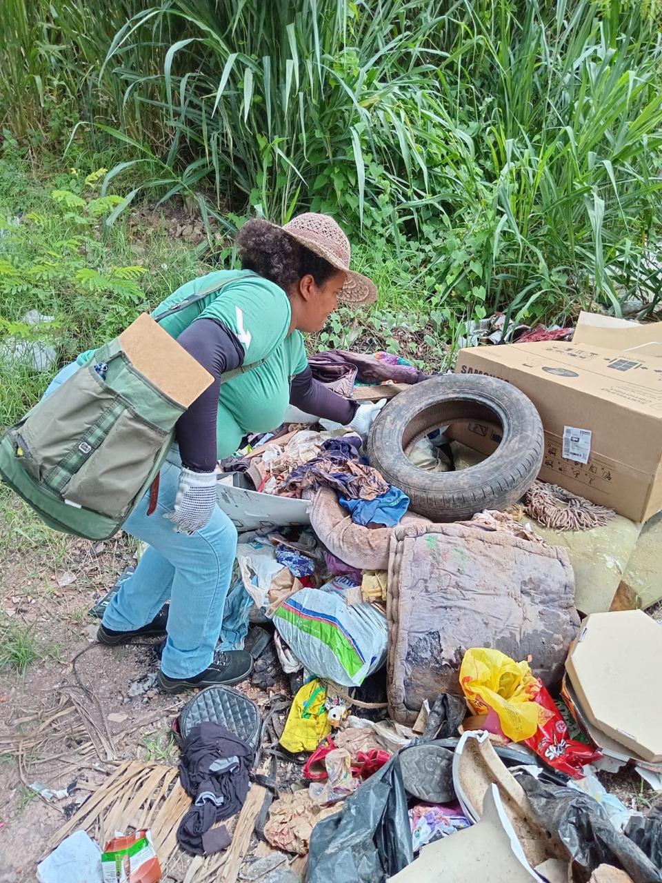 A imagem mostra uma mulher de pele morena, vestindo uma camisa verde e calças azuis, agachada perto de um monte de lixo. Ela está usando um chapéu de palha e uma bolsa verde escura. O lixo inclui vários itens como um pneu de carro, caixas de papelão, roupas, embalagens plásticas e outros detritos. O cenário é exterior, em uma área com vegetação, sugerindo um ambiente urbano ou peri-urbano. A mulher parece estar recolhendo ou inspecionando o lixo, possivelmente como parte de uma atividade de limpeza ou pesquisa. A imagem sugere preocupações com a gestão de resíduos e o impacto ambiental do lixo.