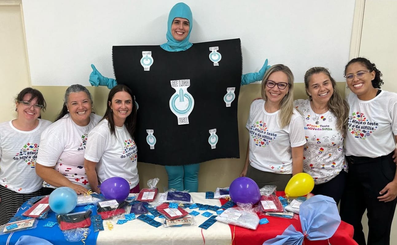 A imagem mostra sete mulheres e uma pessoa usando um figurino de interruptor de energia, todas sorrindo para a câmera. As mulheres estão usando camisetas brancas com mensagens em português sobre o teste de HIV e sífilis. Elas estão posicionadas atrás de uma mesa com preservativos, folhetos e outros materiais informativos sobre saúde sexual. O figurino representa a importância de fazer o teste. A cena parece ser um evento de saúde pública. A foto parece ter sido tirada em um ambiente interno, provavelmente uma escola ou centro comunitário. A atmosfera é alegre e informativa.