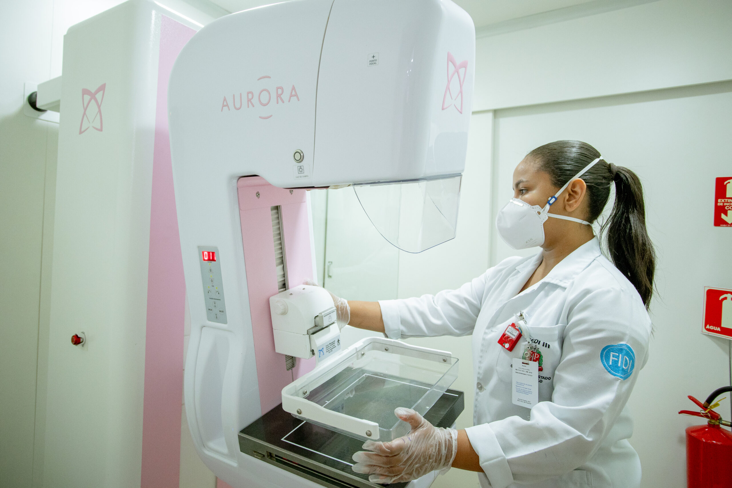A imagem mostra uma profissional de saúde, usando máscara facial e luvas, operando um mamógrafo Aurora. O mamógrafo é de cor rosa e branco, e a profissional de saúde parece estar preparando o equipamento para um exame. O ambiente parece ser uma sala de exames médicos, com paredes brancas e um extintor de incêndio visível ao fundo. A profissional de saúde parece concentrada em seu trabalho. A imagem transmite uma sensação de profissionalismo e cuidado médico.