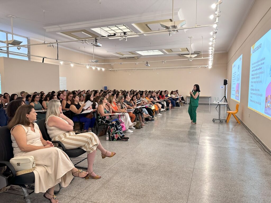 A imagem mostra uma grande sala de conferências ou auditório, com muitas mulheres sentadas em cadeiras dispostas em fileiras.  Uma palestrante está de pé na frente, vestindo um macacão verde e falando para a audiência. Duas telas de projeção mostram apresentações, e um pequeno número de objetos, como mesas e cadeiras, estão visíveis no fundo da sala.