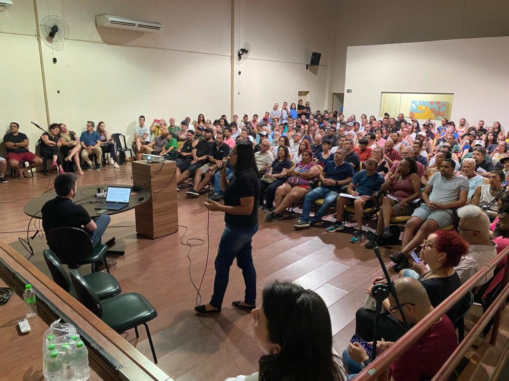 Perspectiva do palco mostrando a palestrante, uma mulher de camisa preta e calça jeans, falando ao público lotado. À sua frente, uma mesa com um notebook e garrafas de água. A plateia acompanha atentamente, com alguns tomando notas. O ambiente tem piso de madeira, ar-condicionado e ventiladores fixados na parede.