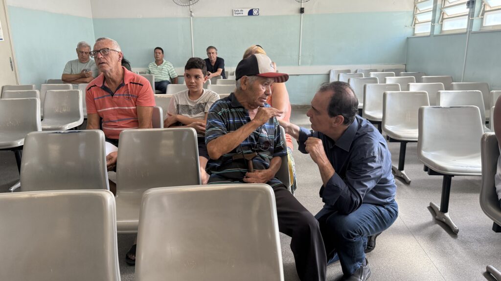 A imagem mostra uma sala de espera de um hospital ou clínica. Há várias cadeiras cinzas de plástico alinhadas em fileiras. Várias pessoas estão sentadas esperando, algumas sozinhas e outras conversando. A maioria das pessoas são homens, de meia-idade a mais velhos. Há uma pessoa mais jovem sentada entre dois adultos mais velhos. Uma pessoa de camisa azul escura está agachada, aparentemente conversando com um homem mais velho que usa um boné e segura um bengala. No fundo, pode-se ver uma placa que indica "Sala de Espera". A iluminação da sala é natural e o ambiente é tranquilo. As paredes são pintadas de um tom pastel claro. Há um ventilador de teto e uma janela com grades. A imagem transmite uma sensação de espera paciente, mas não há indicação de estresse ou ansiedade aparente nas pessoas presentes.