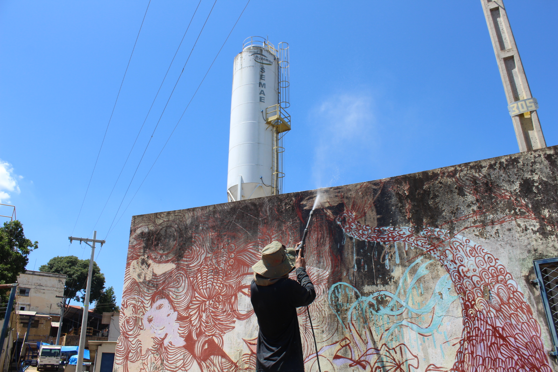 A imagem mostra um homem usando um limpador de alta pressão para limpar uma parede de concreto grande com um grafite. O grafite é uma peça colorida e intrincada de arte de rua, que apresenta uma variedade de formas e cores. O homem está de costas para a câmera e está vestido com um chapéu e roupas escuras. A parede está localizada em uma área urbana, com edifícios e postes de energia visíveis no fundo. Há também um silo branco grande visível ao fundo. O céu está claro e azul. A imagem transmite uma sensação de atividade e renovação, pois o homem está limpando a parede e atualizando a arte de rua.