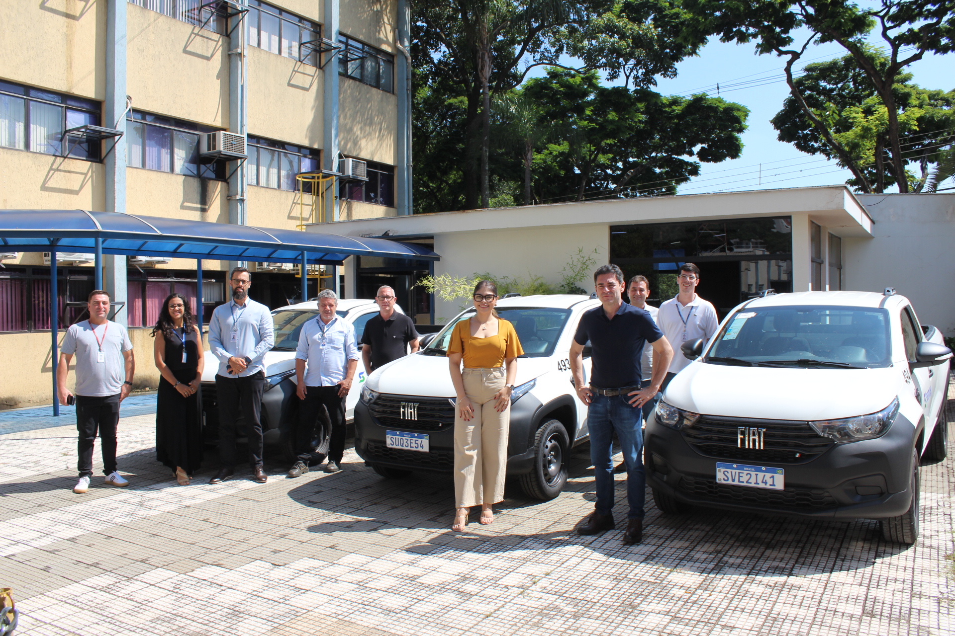 A foto mostra um grupo de pessoas em pé na frente de uma série de caminhonetes Fiat Strada brancas. O grupo inclui homens e mulheres de diferentes idades, todos vestidos com roupas casuais. Eles estão sorrindo para a câmera. As caminhonetes estão estacionadas em uma fileira, e há um prédio atrás deles. O fundo é um pouco desfocado, mas é possível ver árvores e céu. A foto parece ter sido tirada ao ar livre, em um dia ensolarado.