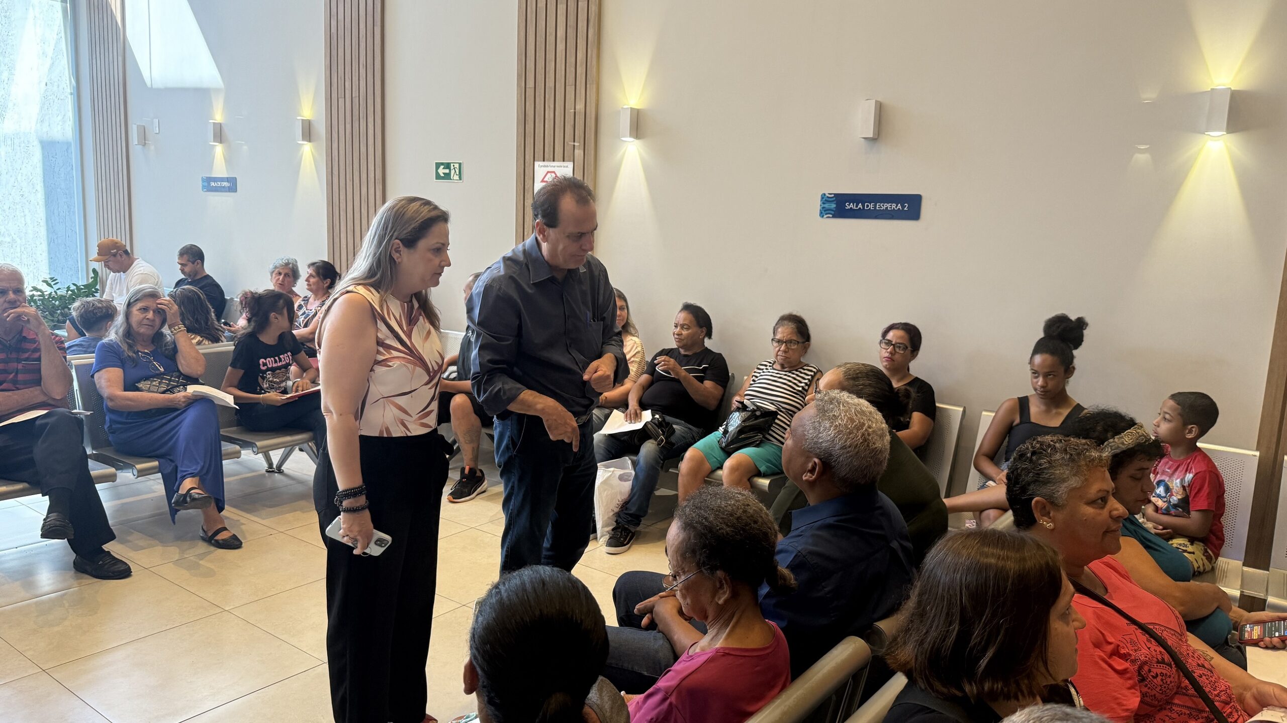 A imagem mostra uma sala de espera lotada em um ambiente que parece ser uma clínica ou hospital. Há muitas pessoas sentadas em cadeiras de espera, algumas individualmente, outras em grupos. As pessoas parecem estar esperando para serem atendidas. Há um casal que parece estar conversando com outras pessoas na sala. A sala é bem iluminada e limpa. Há cartazes na parede com informações em português, incluindo uma placa que indica "Sala de Espera 2". As pessoas na imagem são de diversas idades e etnias. A atmosfera parece calma e organizada, embora esteja lotado. Há também algumas plantas em vasos na sala.