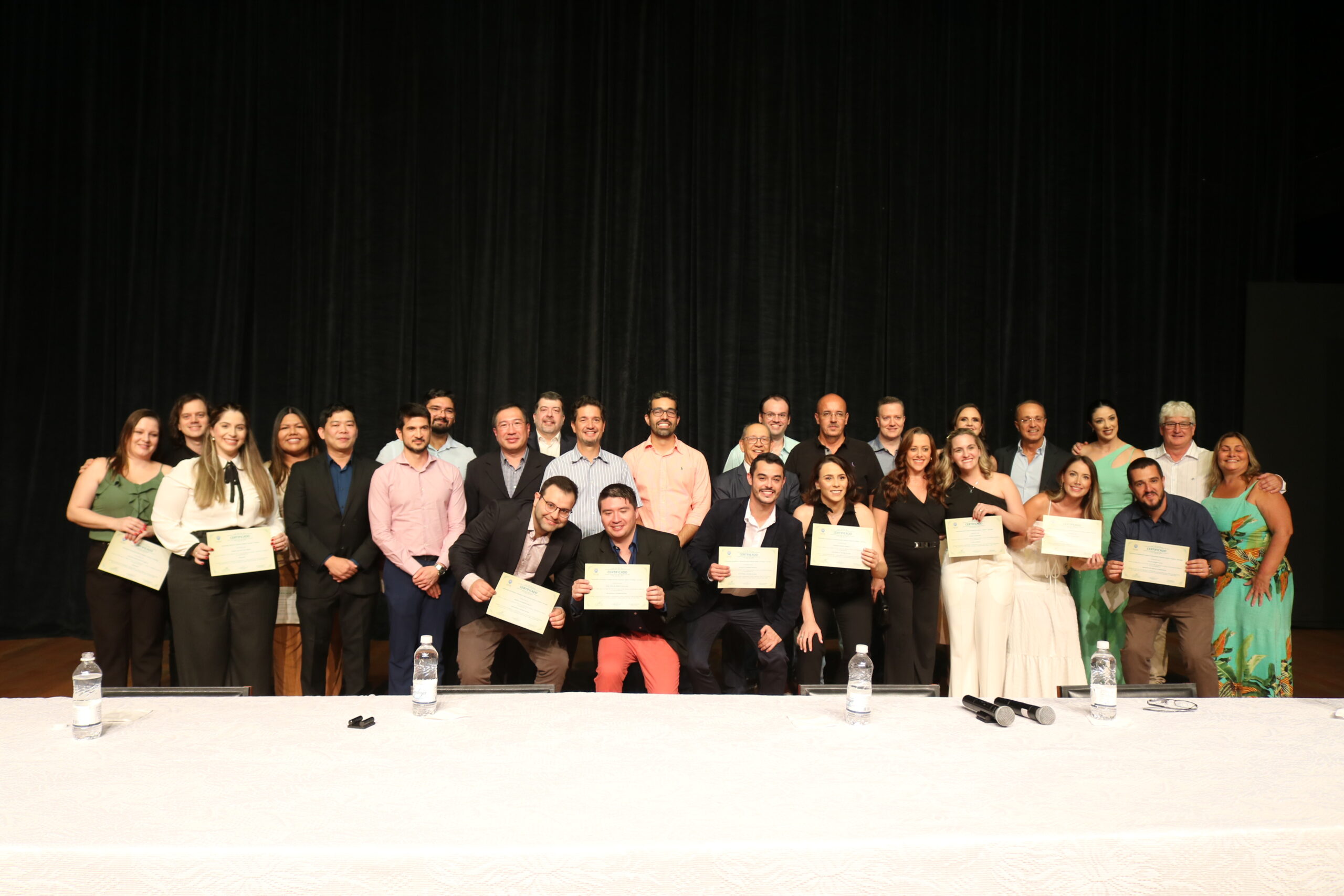 A imagem mostra um grupo de aproximadamente 30 pessoas, homens e mulheres, em pé em duas fileiras na frente de um palco escuro. Eles estão segurando certificados e parecem estar em um evento de formatura ou premiação. As pessoas estão sorrindo e algumas estão abraçadas. Elas estão vestidas com roupas formais e informais. A iluminação é focada no grupo de pessoas, deixando o fundo escuro e pouco visível. Há uma mesa branca na frente do grupo com garrafas de água e microfones.