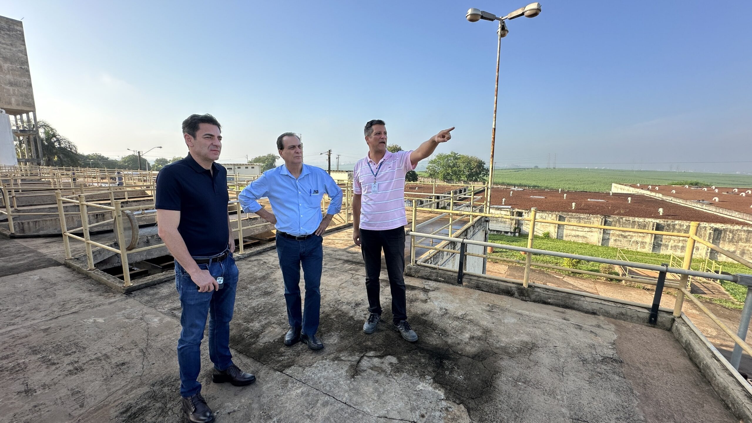 Três homens estão em pé em uma plataforma de concreto ao ar livre, observando uma instalação industrial. O homem da direita está apontando para algo à distância. O homem do meio está de braços cruzados e o homem da esquerda está com as mãos nos bolsos. O céu está claro e o sol está brilhando. A instalação parece ser uma estação de tratamento de água ou uma planta industrial semelhante. Há um campo de cana-de-açúcar ao fundo.