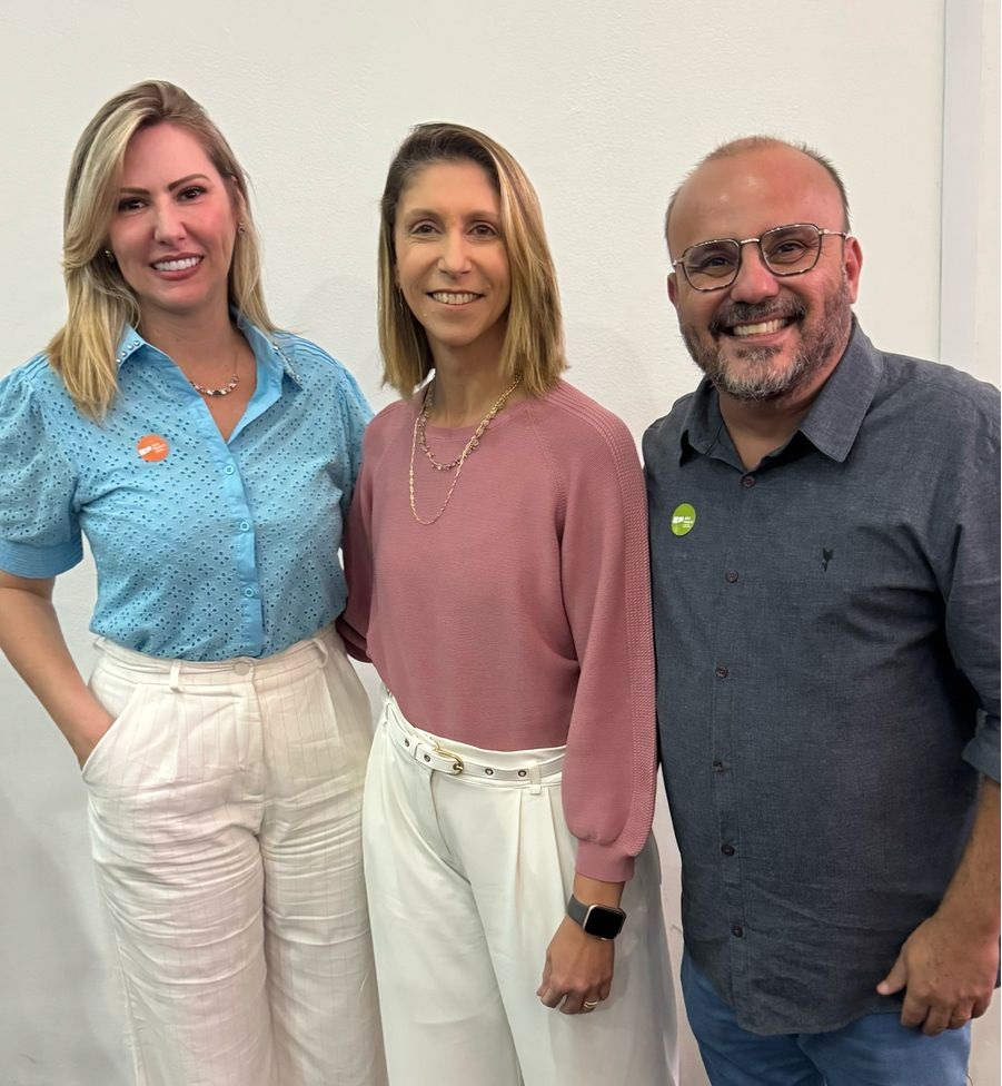 Três pessoas posam sorrindo para a foto contra um fundo branco. À esquerda, uma mulher de blusa azul clara e calça branca. No centro, outra mulher de blusa rosa e calça branca. À direita, um homem de camisa azul escura e jeans. Eles estão próximos, demonstrando entrosamento.