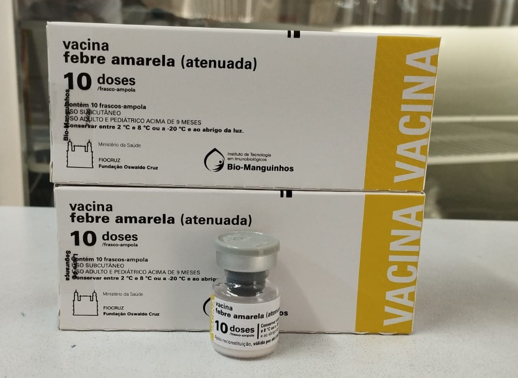 A imagem mostra duas caixas de vacina contra febre amarela empilhadas, com uma ampola de vacina na frente. As caixas são brancas com detalhes amarelos e texto em português. O texto indica que cada caixa contém 10 doses da vacina, que deve ser armazenada em temperaturas específicas. Há também informações sobre o fabricante (Bio-Manguinhos) e os órgãos responsáveis pela vacina. A ampola é pequena e transparente, com um tampão cinza. A imagem é bem iluminada e os detalhes são nítidos.