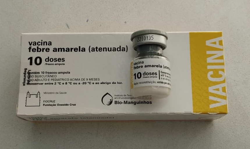 A imagem mostra uma caixa de vacina contra a febre amarela. A caixa é branca e amarela, com o nome da vacina e outras informações impressas nela. A caixa contém 10 doses de vacina. A vacina é produzida pela Bio-Manguinhos, um instituto de tecnologia em imunobiológicos. A imagem também mostra um frasco de vacina. O frasco é de vidro e contém um pó branco. O frasco está rotulado com o nome da vacina e outras informações.