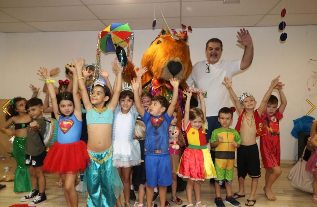 Crianças fantasiadas em uma festa de Carnaval posam animadas com os braços para cima, ao lado de um homem de camisa branca e uma mascote vestida com um grande traje de urso marrom decorado com fitas coloridas. O ambiente fechado tem decoração festiva, com um guarda-chuva de frevo no alto.
