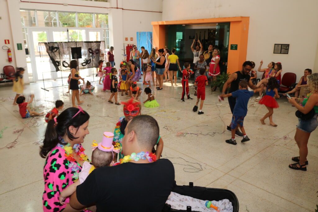 Salão amplo e iluminado com crianças e adultos participando de uma festa de Carnaval. Algumas crianças brincam no chão com serpentinas, enquanto outras dançam e interagem. No primeiro plano, um casal fantasiado cuida de um bebê.