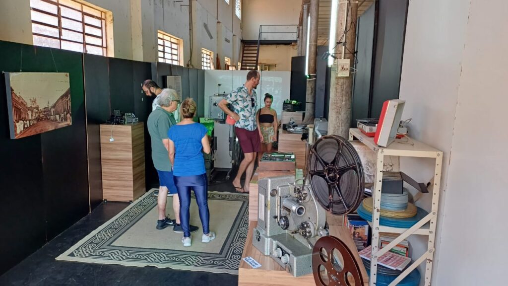 Visitantes exploram uma exposição de equipamentos de cinema e audiovisual em um espaço rústico, com paredes altas, janelas com grades e iluminação suave. No primeiro plano, um projetor de filme antigo com rolos metálicos está exposto sobre um balcão de madeira. Algumas pessoas observam e interagem com as peças, enquanto no fundo há um telão exibindo imagens históricas.