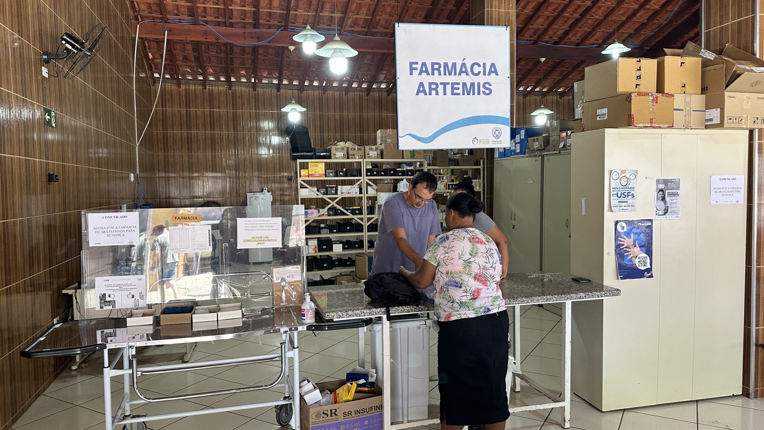 A imagem mostra o interior de uma farmácia chamada "Farmácia Artemis". Há várias prateleiras cheias de medicamentos e outros itens farmacêuticos. Duas mulheres estão na frente do balcão, aparentemente atendendo clientes, e um homem está próximo a elas, talvez um farmacêutico ou funcionário. Há também uma quantidade significativa de caixas empilhadas em um armário, sugerindo estoque ou remessas recentes. Há avisos afixados na parede, um deles informando sobre um novo horário e outro sobre mudança de local da farmácia. O ambiente parece ser simples, típico de uma farmácia em uma região mais rural ou com recursos limitados. Um carrinho de transporte médico está visível perto do balcão, o que pode indicar que a farmácia está integrada a um centro de saúde ou serviço médico. A iluminação é feita por lâmpadas penduradas no teto. A decoração é simples, com paredes revestidas de madeira.