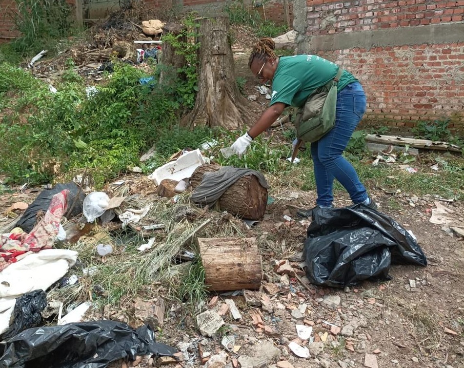 A imagem mostra uma mulher vestindo luvas brancas e uma camiseta verde, recolhendo lixo em um terreno baldio. O local está repleto de detritos, incluindo pedaços de madeira, sacos plásticos pretos, roupas velhas, e outros resíduos diversos. A vegetação rasteira e alguns troncos de árvores são visíveis no cenário. Ao fundo, há uma parede de tijolos. A mulher parece estar envolvida em uma atividade de limpeza ou remoção de lixo do local. A cena sugere um ambiente urbano degradado que necessita de intervenção para a limpeza e recuperação ambiental.