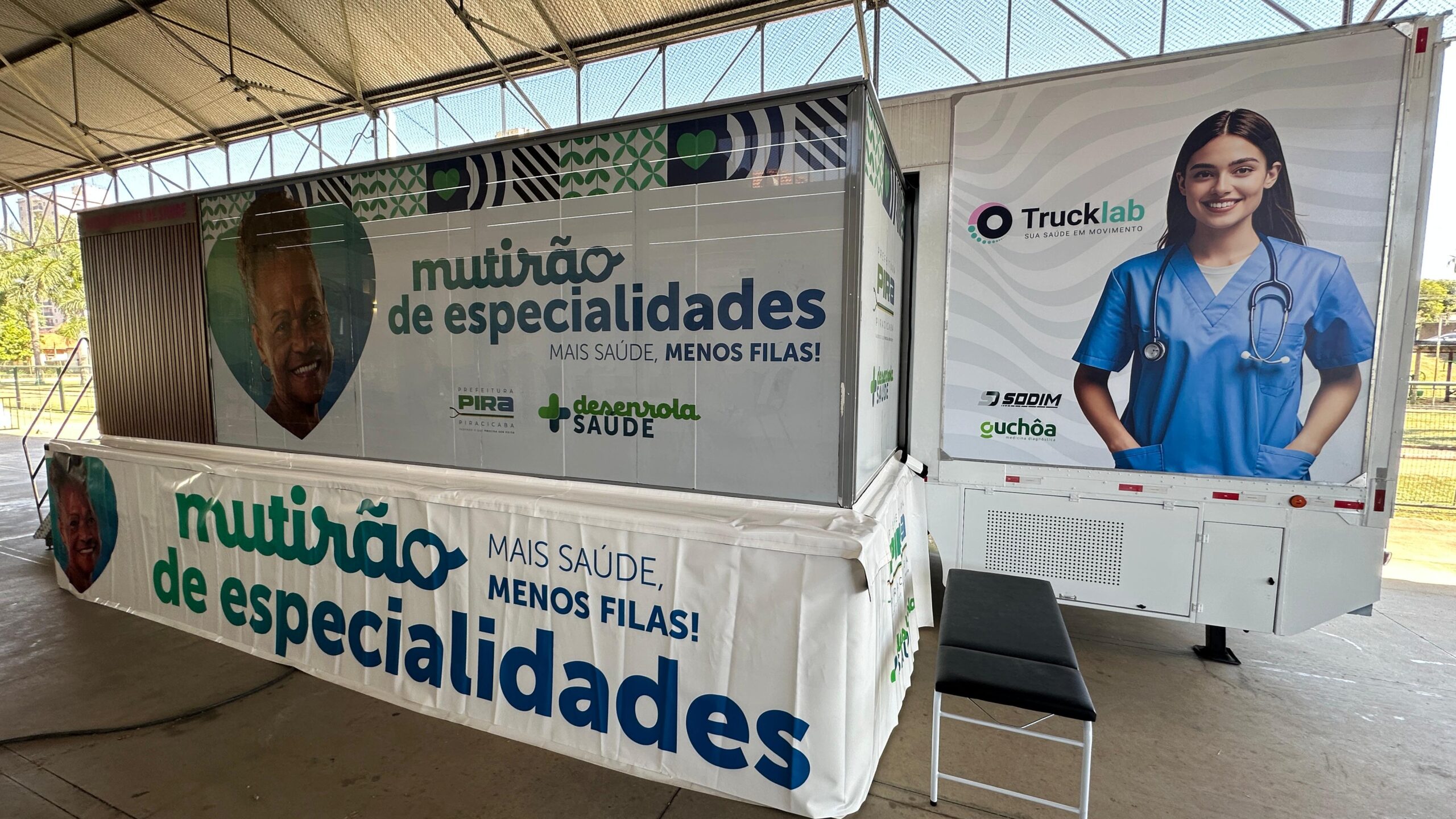 A imagem mostra um caminhão adaptado como unidade móvel de saúde, estacionado em uma área externa coberta. Há grandes faixas em português anunciando um "mutirão de especialidades", com a frase "Mais saúde, menos filas!" repetida em várias partes da estrutura. *Detalhes da imagem:* * *O Caminhão:* Um caminhão branco, com a marca "Trucklab" visível, indicando que se trata de uma unidade móvel. Na lateral, há uma imagem de uma enfermeira ou médica, e logotipos de patrocinadores ou parceiros, como "SODIM" e "Guchôa". A parte traseira do caminhão tem uma porta e uma grade de ventilação. * *As Faixas:* Grandes faixas, com fundo claro e letras em azul e verde, anunciam o "Mutirão de Especialidades". A repetição da frase "Mais saúde, menos filas!" destaca o objetivo da iniciativa. Há também os logotipos da prefeitura de Piracicaba e da iniciativa "Desenrola Saúde". As faixas estão em português. * *O Local:* O caminhão está estacionado em um local que parece ser um pátio ou área coberta, possivelmente um espaço público destinado a eventos. Há uma estrutura de cobertura metálica visível acima do caminhão. * *Público-alvo:* A imagem sugere que o mutirão é direcionado à população em geral, com foco na melhoria do acesso a serviços de saúde especializados e na redução do tempo de espera. *Em resumo:* A imagem documenta um evento de saúde pública, um "mutirão de especialidades médicas", utilizando uma unidade móvel para facilitar o acesso a serviços de saúde, especialmente em um local onde há filas significativas. A iniciativa parece ser uma parceria entre a prefeitura de Piracicaba, outras organizações e empresas privadas.