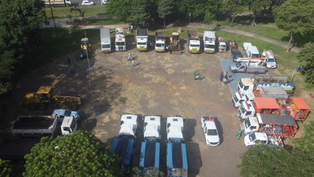 A imagem mostra uma visão aérea de um estacionamento ou área aberta organizada em uma formação semi-circular. Há vários tipos de veículos e equipamentos, incluindo caminhões, veículos utilitários e máquinas de construção. Algumas pessoas estão visíveis, algumas paradas próximas aos veículos e outras caminhando pela área. O local é cercado por vegetação, com árvores e um caminho ao fundo. A cena parece organizada, possivelmente para um evento, exibição ou reunião relacionada a serviços municipais ou de construção.