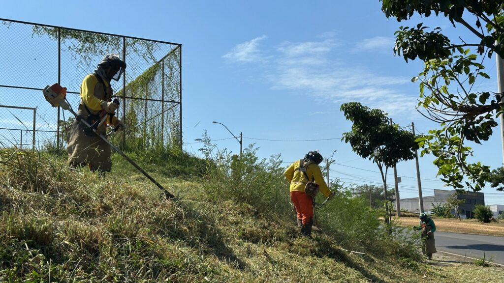 A imagem mostra três pessoas realizando trabalho de paisagismo ou manutenção em uma encosta coberta de grama. Elas estão utilizando roçadeiras para aparar a vegetação. Os trabalhadores usam equipamentos de proteção, como capacetes e viseiras, para garantir a segurança durante o serviço. Ao fundo, há uma cerca de arame com vegetação crescendo ao redor, além de um céu azul claro.