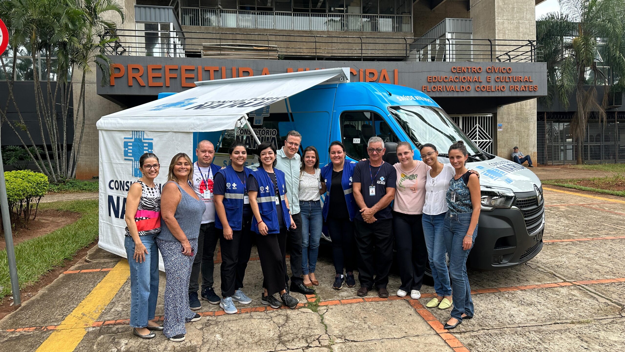 A imagem mostra um grupo de pessoas em frente a uma van adaptada como consultório móvel, estacionada em frente ao Centro Cívico Educacional e Cultural Florivaldo Coelho Prates. *Detalhes da imagem:* * *Local:* A foto foi tirada em frente a um prédio identificado como "PREFEITURA MUNICIPAL" (provavelmente a prefeitura da cidade) e "CENTRO CÍVICO EDUCACIONAL E CULTURAL FLORIVALDO COELHO PRATES". Isso indica um ambiente público e institucional. * *Veículo:* Uma van branca e azul, com a inscrição "CONSULTÓRIO MÓVEL" e um logotipo de cruz médica, está claramente identificada como uma unidade móvel de saúde. A van parece bem equipada e preparada para atendimento médico. * *Pessoas:* Um grupo de aproximadamente doze pessoas, a maioria mulheres, posam para a foto em frente à van. Elas parecem ser profissionais de saúde ou funcionários ligados à iniciativa do consultório móvel, considerando suas vestimentas e a proximidade com a van. Algumas usam uniformes ou jalecos. Há uma diversidade de idades e aparências no grupo. * *Contexto:* A cena sugere uma iniciativa de saúde pública, possivelmente um programa de atendimento médico em locais de difícil acesso ou com pouca infraestrutura, utilizando uma unidade móvel para levar os serviços de saúde à comunidade. A presença da prefeitura reforça a ideia de um projeto governamental ou com apoio público. * *Outras observações:* O ambiente parece limpo e organizado. Há um pequeno toldo ou tenda anexa à van, possivelmente para ampliar o espaço de atendimento. No fundo, é possível ver um pouco da vegetação e parte de um estacionamento. Uma pessoa está sentada em um banco ao fundo, observando a cena. Em resumo, a imagem documenta um evento relacionado a uma iniciativa de saúde pública, utilizando um consultório móvel para levar atendimento à comunidade, possivelmente em uma cidade brasileira. A imagem captura um momento de interação entre os profissionais de saúde envolvidos no projeto.