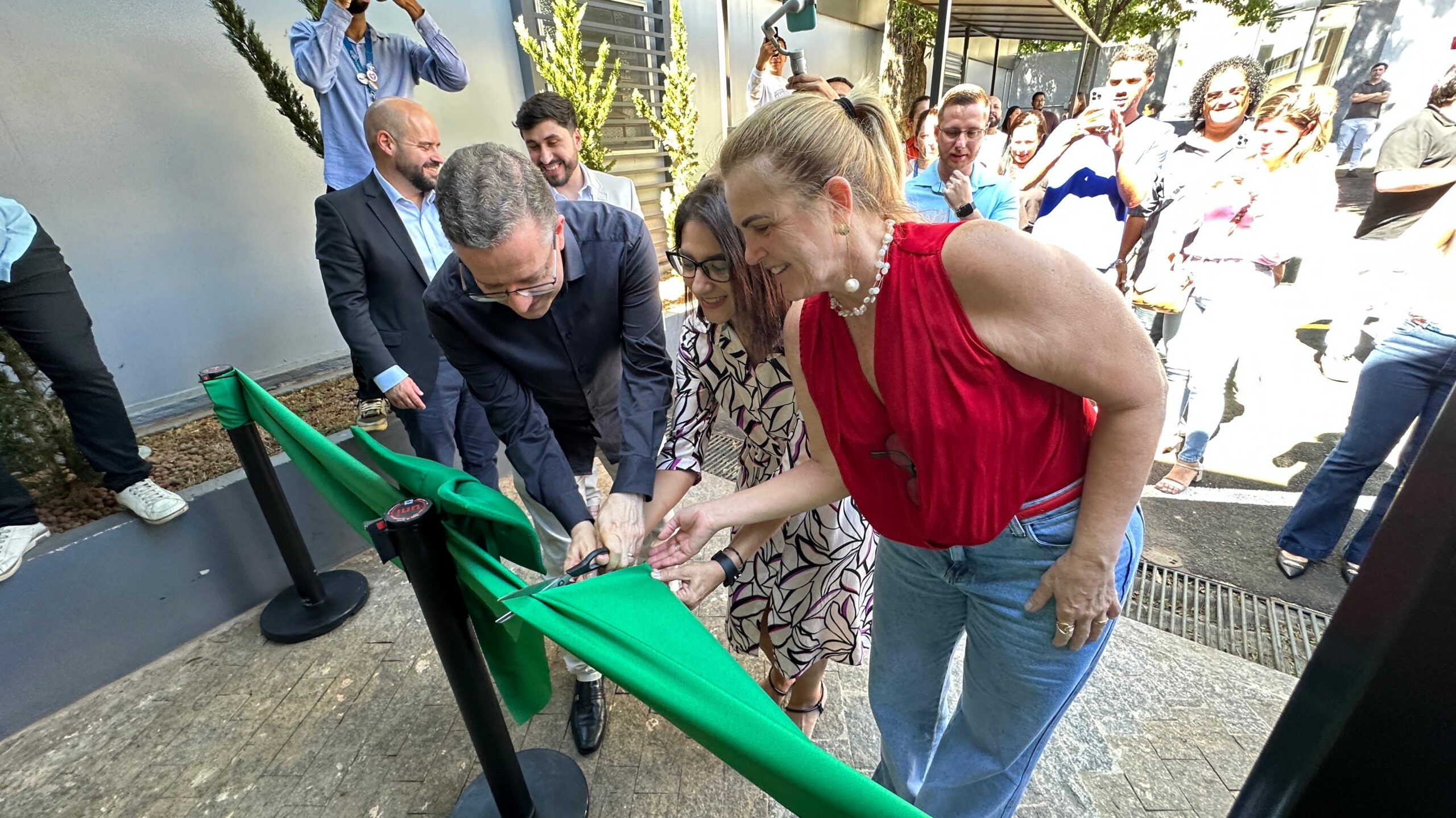 A imagem mostra um corte de fita inaugural, provavelmente de um novo estabelecimento ou projeto. Várias pessoas estão presentes, participando do evento. No centro da imagem, duas mulheres estão cortando uma fita verde com uma tesoura. Uma delas veste um vestido vermelho e a outra um vestido com estampa floral. Um homem está ao lado delas, ajudando no corte da fita. Ao fundo, outras pessoas assistem à cerimônia, algumas tirando fotos. Há um grupo maior de pessoas à direita, e outras menos numerosas à esquerda. O ambiente parece ser externo, com uma área pavimentada e alguns elementos arquitetônicos visíveis. A atmosfera geral é de celebração e formalidade. A imagem sugere um evento público importante, possivelmente uma inauguração oficial.