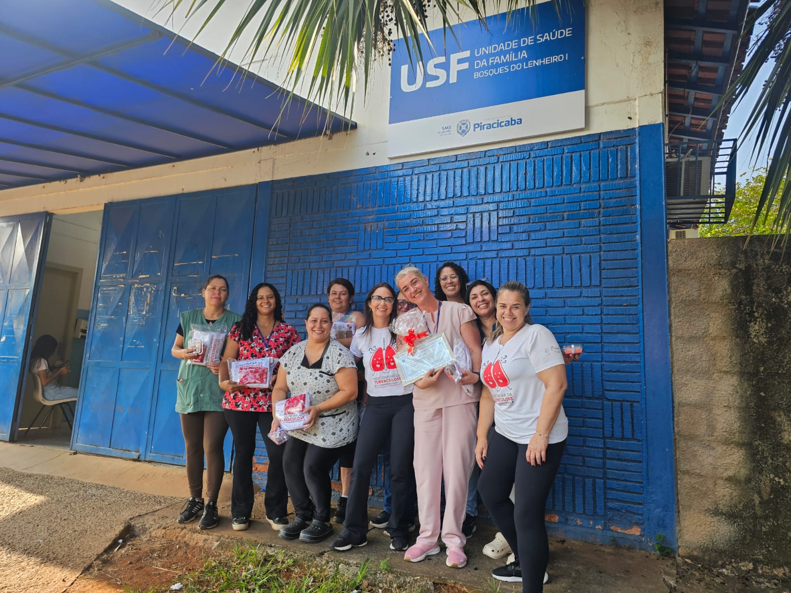 A imagem mostra um grupo de mulheres em frente a uma Unidade de Saúde da Família (USF). A USF é identificada como "USF Bosques do Lenheiro I" e pertence à prefeitura de Piracicaba. As mulheres estão sorrindo para a câmera e segurando vários itens, incluindo o que parecem ser pacotes de presentes ou materiais de conscientização. Uma das mulheres está segurando um certificado ou placa de agradecimento. Elas estão vestidas com roupas casuais, algumas com camisetas com o logo de uma campanha contra a tuberculose. A parede do edifício é azul. A cena sugere uma comemoração ou evento relacionado à saúde, possivelmente um reconhecimento do trabalho da equipe ou uma campanha de conscientização sobre a tuberculose. A atmosfera é positiva e colaborativa.