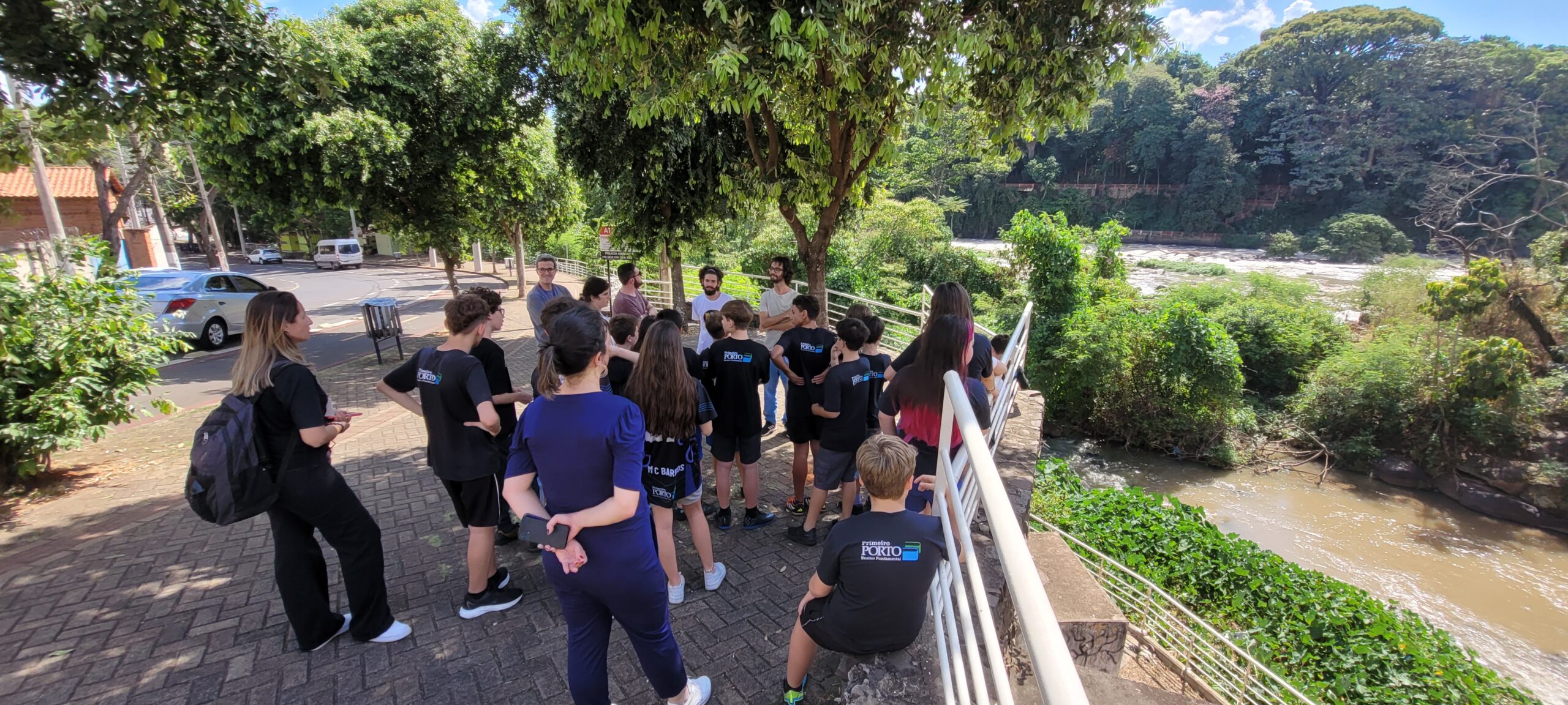 A imagem mostra um grupo de estudantes e professores em uma excursão perto de um rio. Eles estão parados em uma calçada ao lado de uma cerca branca, observando o rio que flui abaixo. Há árvores e vegetação exuberante ao redor do rio. Os estudantes parecem estar ouvindo atentamente um professor ou guia que está falando com eles. Há alguns carros estacionados na rua atrás deles. O ambiente parece ser um local público, talvez um parque ou área de preservação ambiental. O grupo parece estar participando de uma atividade educacional ou de uma visita guiada. A imagem sugere um dia ensolarado e agradável. O clima parece ser tropical ou subtropical, considerando a vegetação. Pelo menos uma das pessoas na foto usa uma camiseta com a palavra "PORTO", possivelmente indicando uma escola, organização ou o nome do local.