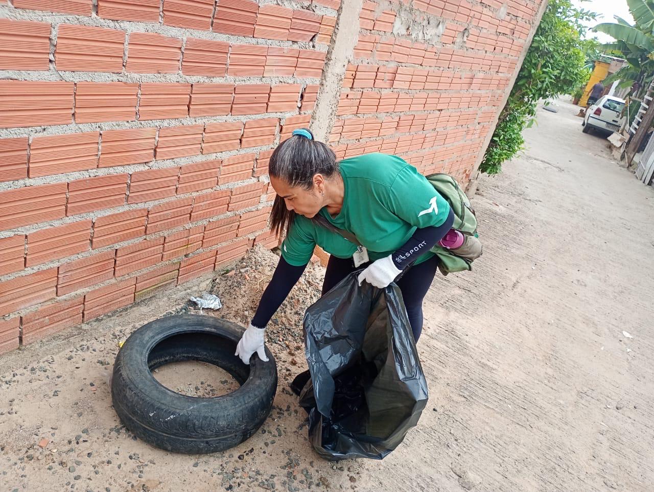 A imagem mostra uma mulher de camisa verde e luvas brancas recolhendo um pneu usado em uma rua de chão batido ao lado de um muro de tijolos. Ela está colocando o pneu em um saco de lixo preto grande. Ela carrega uma mochila verde escura com uma garrafa de água roxa visível. Ao fundo, há uma rua estreita com casas e um carro estacionado. A cena sugere uma ação de limpeza ou trabalho voluntário em uma área urbana. A iluminação indica que é dia.