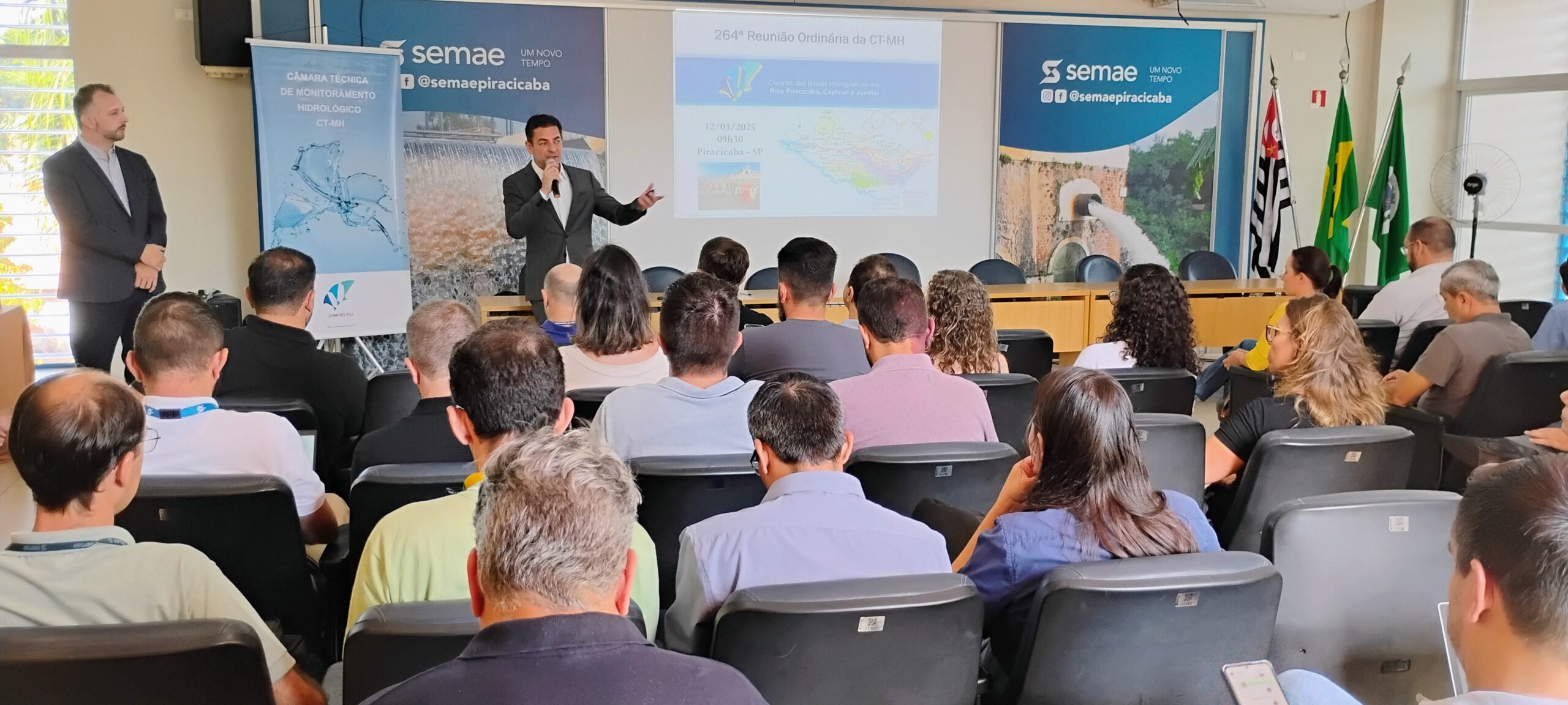 Esta imagem mostra uma apresentação em uma sala de conferências. Um homem está dando uma apresentação usando um projetor. A apresentação está em português e parece ser sobre monitoramento hidrológico. Há várias pessoas na plateia, sentadas em cadeiras. Há também cartazes com a logo da Semae (provavelmente uma empresa ou organização de serviços de água e esgoto) e informações sobre a reunião. As bandeiras do Brasil e do estado de São Paulo estão visíveis no fundo. A imagem sugere uma reunião formal ou apresentação técnica, possivelmente um encontro da Câmara Técnica de Monitoramento Hidrológico (CT-MH) da Semae de Piracicaba. O número 264ª indica que esta é a 264ª reunião ordinária desse comitê. A apresentação se concentra em dados cartográficos e informações sobre os rios Piracicaba, Capivari e Jundiaí. A presença de participantes e a formalidade do local indicam um evento importante para a organização.