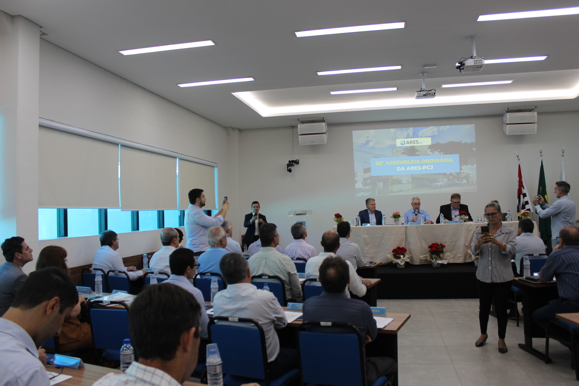 A imagem mostra uma sala de conferências onde está ocorrendo uma assembleia. Há uma tela de projeção exibindo "28ª Assembleia Ordinária da ARES-PC". Várias pessoas estão sentadas em mesas em forma de U, voltadas para uma mesa de palestrantes onde estão sentados quatro indivíduos. Duas pessoas na plateia estão tirando fotos com seus telefones celulares. Há bandeiras brasileiras e outras bandeiras penduradas na parede atrás da mesa de palestrantes. O ambiente demonstra ser um evento formal, possivelmente corporativo ou governamental. A iluminação é artificial, e o ambiente parece limpo e organizado. As pessoas na imagem estão vestidas de forma profissional. A imagem como um todo sugere uma reunião formal de negócios ou uma assembleia de alguma organização.