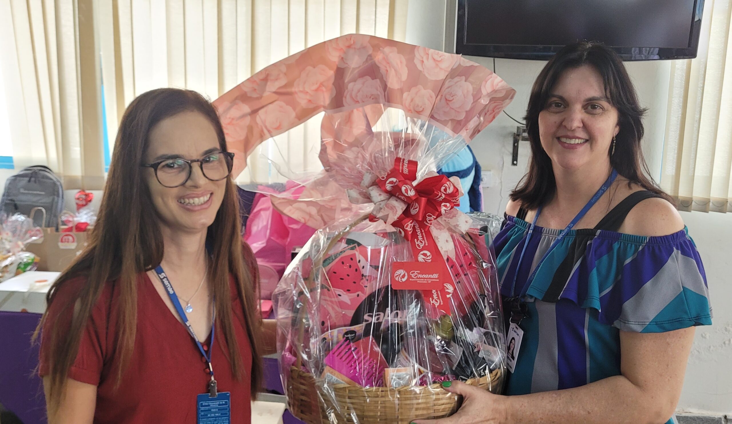 A imagem mostra duas mulheres sorrindo, uma segurando uma cesta de presente grande e elaboradamente embrulhada. A mulher à esquerda está usando óculos de armação escura e uma blusa vermelha. Ela tem cabelos castanhos longos e soltos. A mulher à direita está usando um top sem ombros com listras azuis, cinzas e roxas, e tem cabelos castanhos mais escuros, também longos e soltos. Ela parece mais velha que a outra mulher. A cesta de presente é grande e está cheia de diversos itens, incluindo o que parece ser um conjunto de escova de cabelo, uma bolsa estampada com melancia, e outros itens menores. A cesta está coberta com plástico transparente e um tecido rosa claro com um padrão de rosas. Um laço grande e vermelho está amarrado no topo da cesta. Há uma etiqueta visível com o nome de uma marca, possivelmente "Encanto". O fundo é desfocado, mas mostra um ambiente interno, possivelmente um escritório ou sala de aula, com cortinas na parede e uma televisão na parede ao fundo. Há também algumas sacolas de presente e uma mochila no fundo à esquerda. A iluminação é suave e parece natural. A atmosfera geral da imagem é alegre e festiva, sugerindo uma ocasião de comemoração ou premiação.