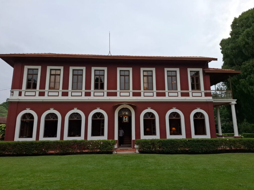 Fachada da Fazenda Dona Catarina, uma construção histórica de dois andares, pintada de vermelho com detalhes brancos e janelas de madeira. Em frente, um gramado bem cuidado e arbustos aparados. A arquitetura é do século XIX, com estilo colonial.